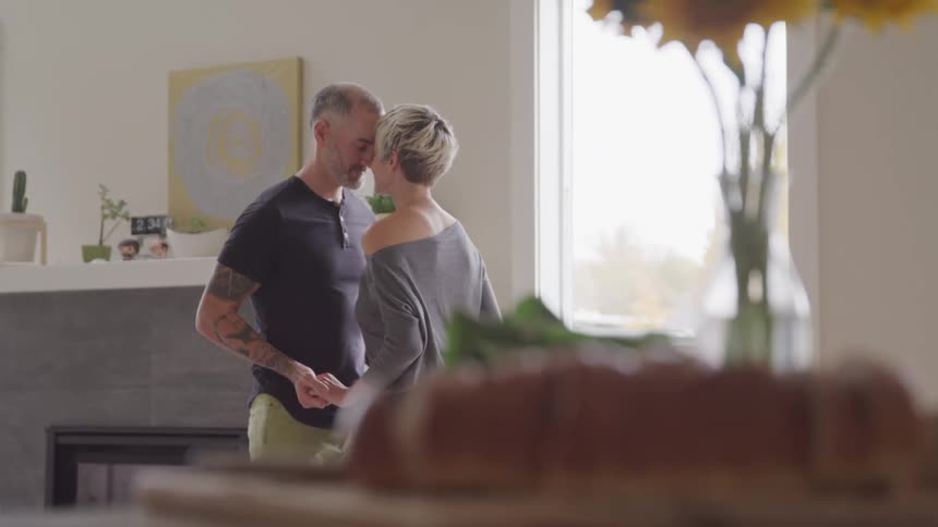A husband and wife hold hands and touch their foreheads together while standing in their living room