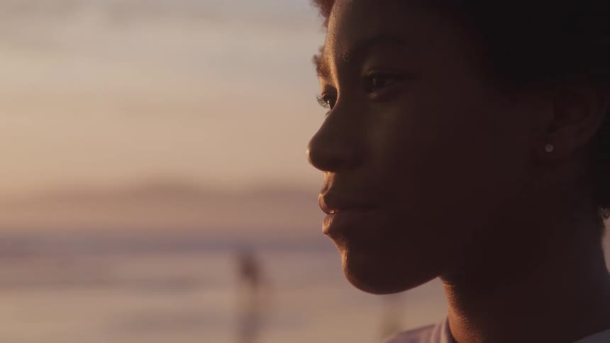 Side view of a black woman standing on the wet sand beach at sunset