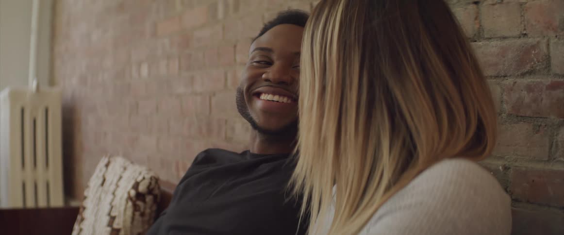 Black man and white woman sitting on a sofa have a talk and laugh
