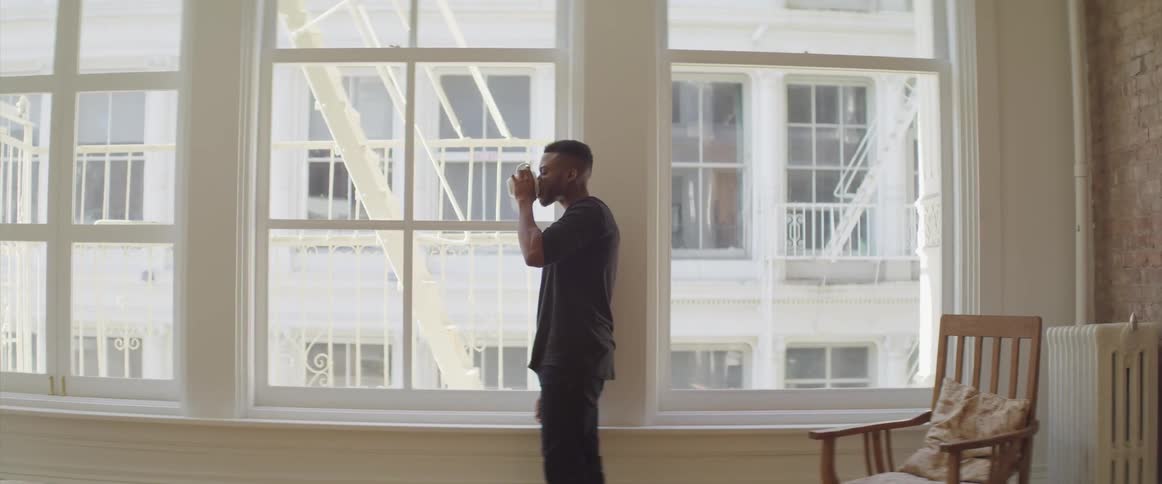 A man takes a drink from a cup standing next to a large window
