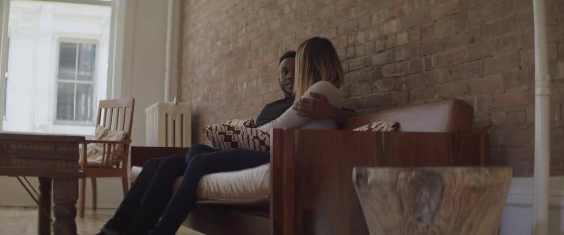 Male has his arm wrapped around female as she sits next to him on the sofa and the talk while looking into each others eyes