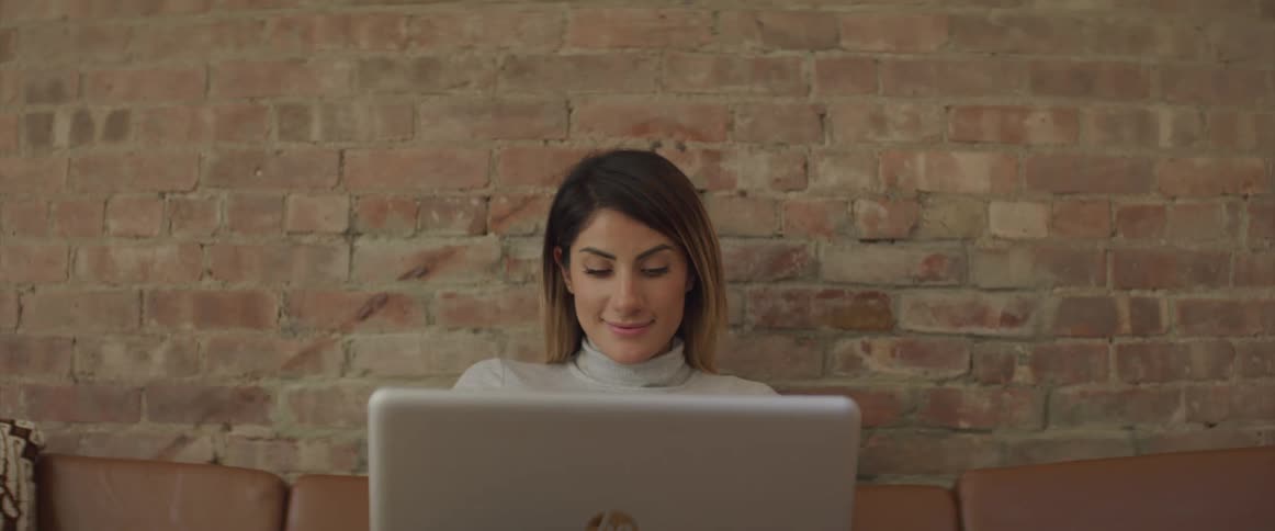 Female sitting on couch against an interior brick wall while looking at her laptop and smiling