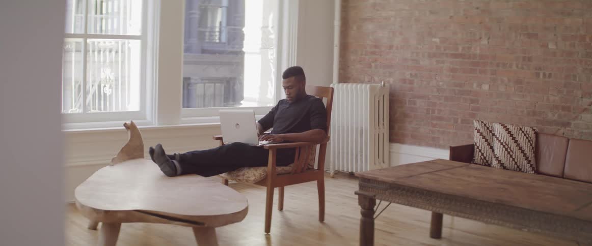 A man sits in a chair in his living room and works on a laptop