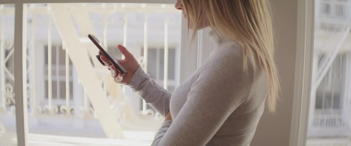 A woman stands in front of a window and scrolls through her phone
