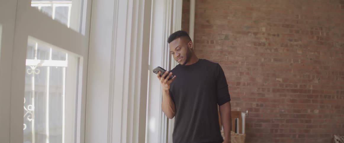 A man smiles as he stands next to his apartment window and uses his smart phone