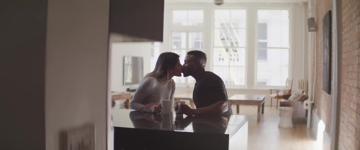 Black man and brunette woman sitting at the table hold cups and kiss each other