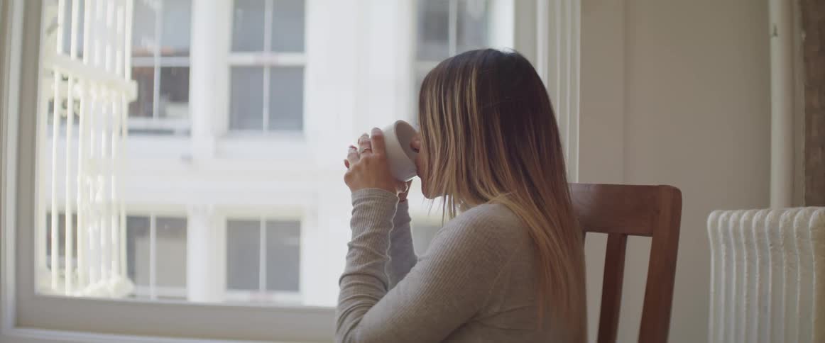 A woman sitting in a chair before a window drinking coffee
