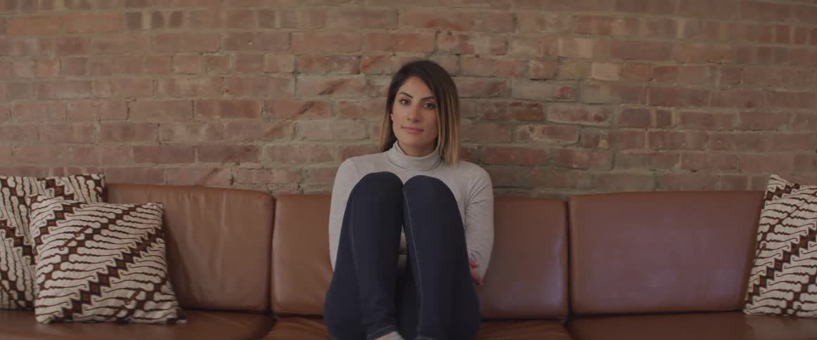 A woman sits by herself on a leather couch against a brick wall