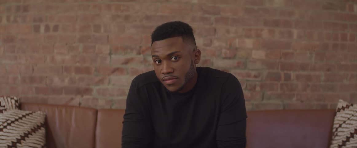 Black man sitting on the couch and looking sideways turns his head to the camera in front of the brick wall