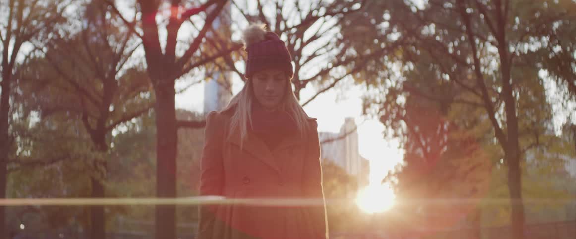 A woman stands still wearing a coat and hat in the park