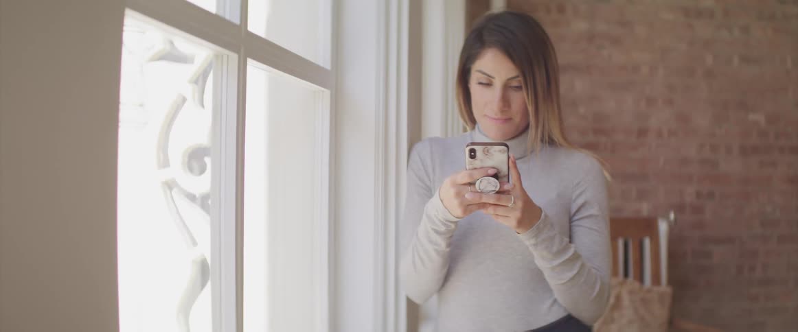 A woman stands beside a window looking at her cell phone