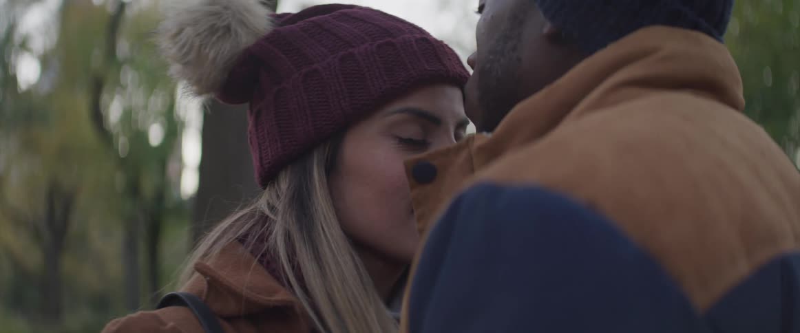 Male and female couple stand facing one another outdoors among the trees in the fall and the female closes her eyes and leans towards the male as he kisses her forehead