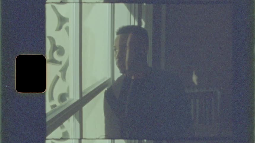 Man leans against window looking out in shadowed room