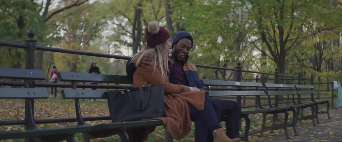 A couple sit together on a park bench with the man having a large smile