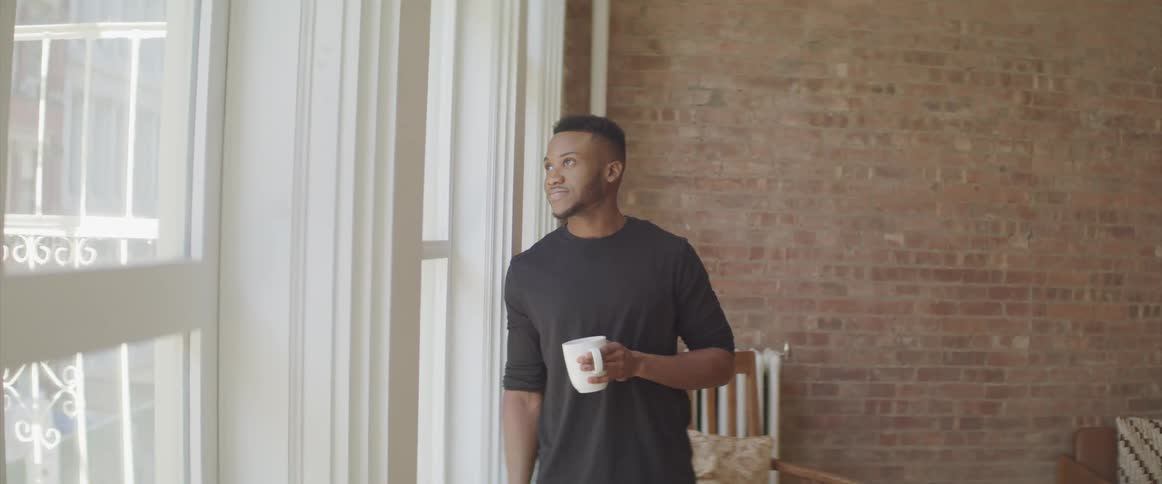 Black man leaning next to window holding coffee mug and looking out