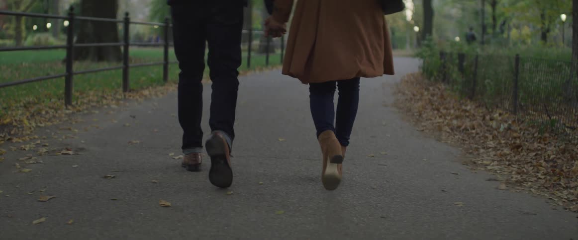 Man and woman walk through a park on a path near a fence and piles of leaves