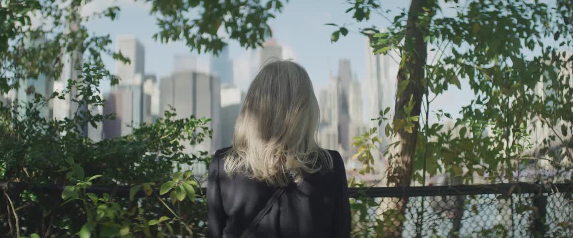A woman standing outdoors looking across a body of water at the skyscrapers of a metropolis