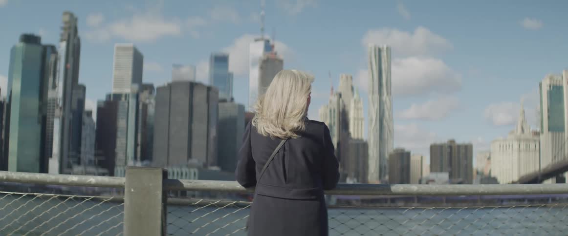 Woman stands at chain link barrier looking at water and downtown on opposite shore