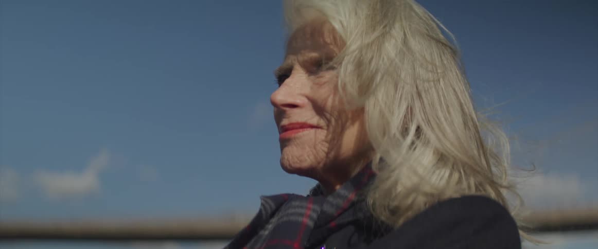 Older woman with ash blonde hair stands under slightly cloudy sky as wind ruffles her hair