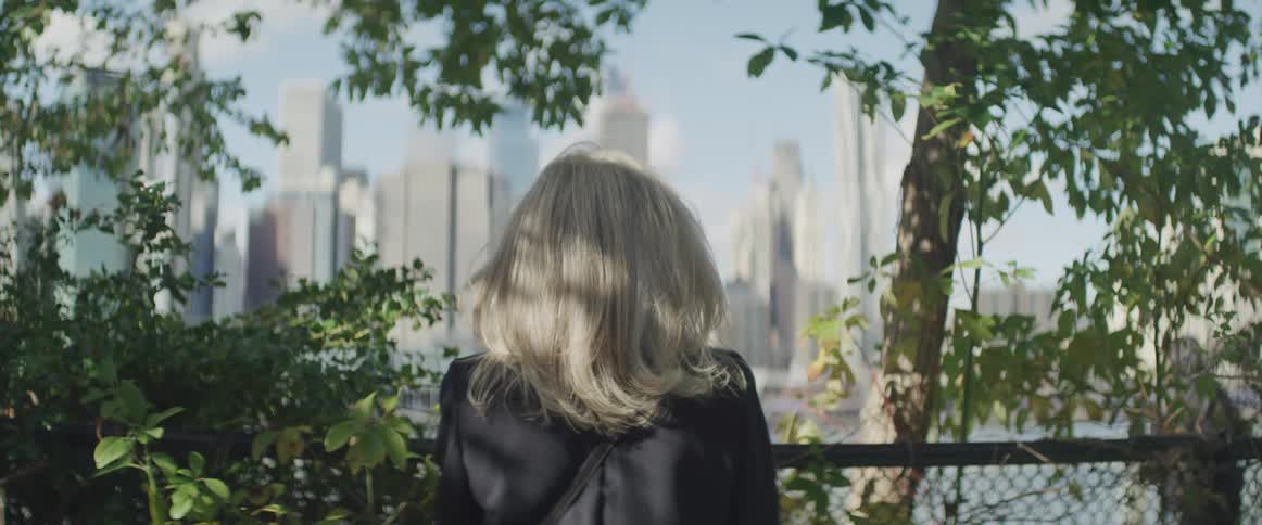 Female stands against a fence along the trees and looks across the river to the city and buildings in the distance