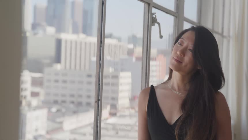 A woman with long hair wearing a sleeveless dress leans against the window in her city apartment on a sunny day
