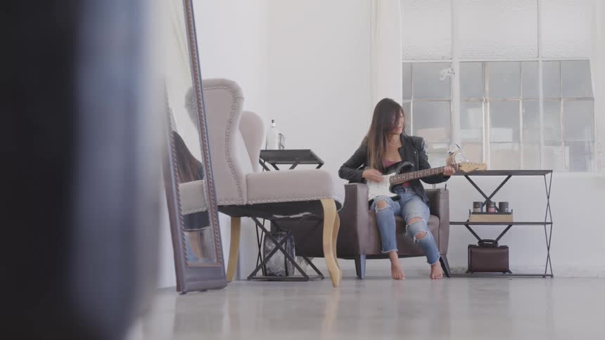 Woman with long dark guitar sitting on an armchair playing a white electric guitar