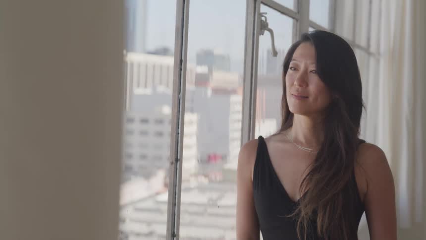 Asian woman looks out the window and smiles slightly in her apartment