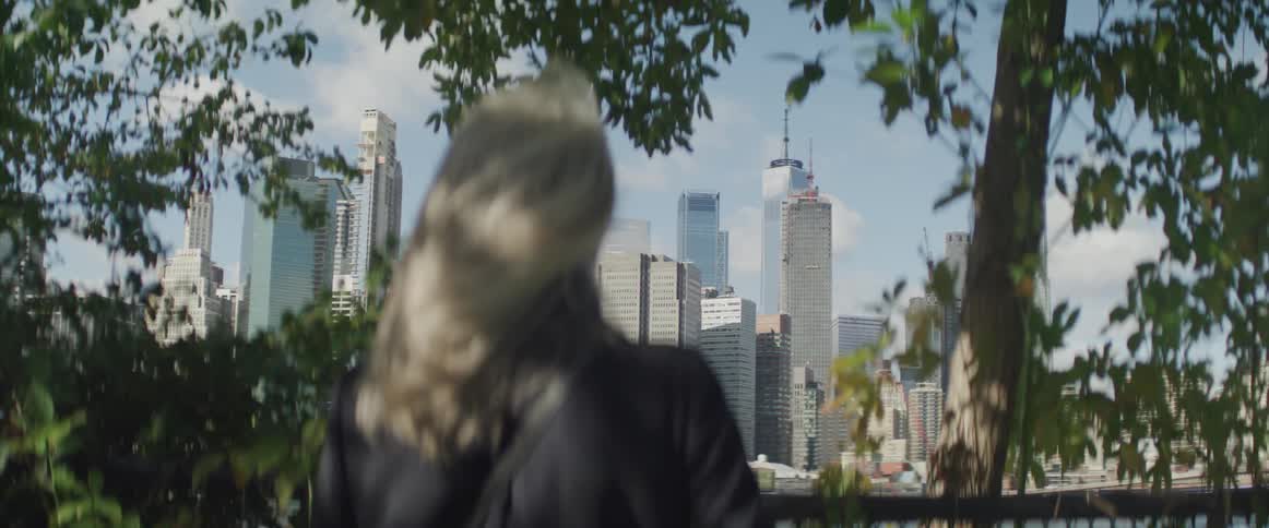 Female standing against a fence peering through the trees at the city skyline across the river