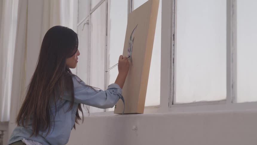 Woman with long dark hair painting on a canvas leaned up to a window indoors
