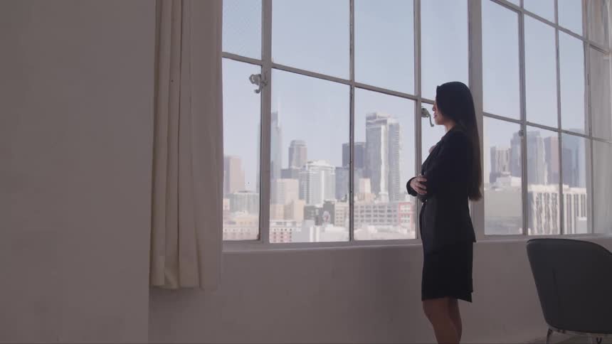 Woman in business attire looking out office window with skyline in view