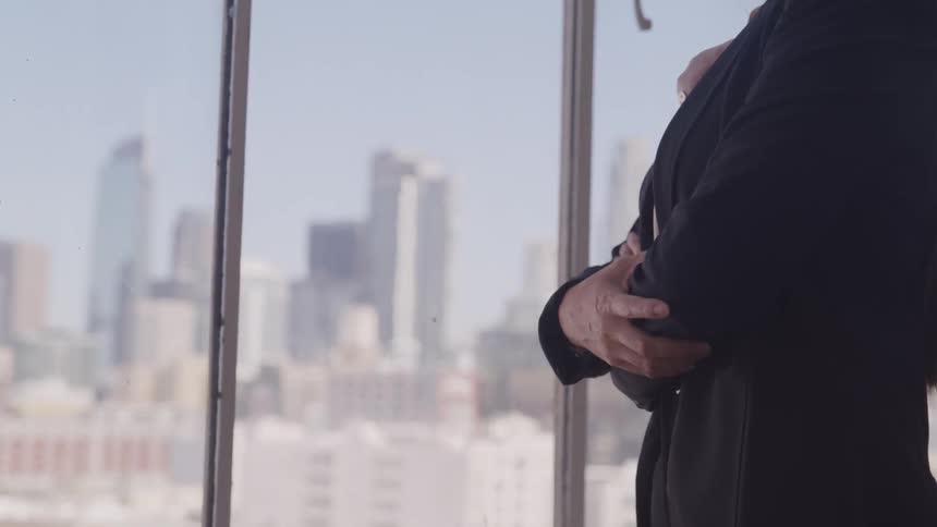 A woman in a business suit stands with arms crossed near large office windows overlooking the city