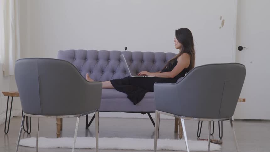 A woman sits with her feet up on a small sofa working on her laptop in a minimalist apartment living room
