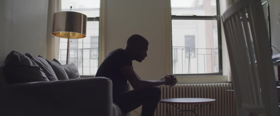A man sitting on a sofa in an apartment with his hands clasped in front of him