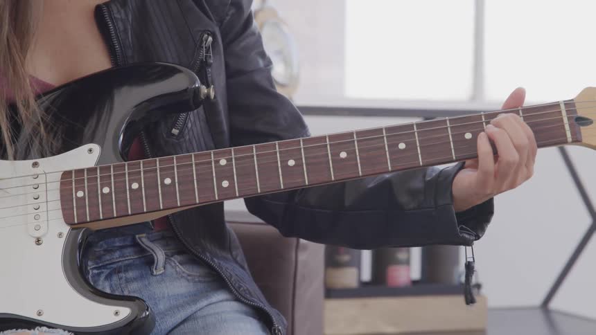 Woman sits in a chair and plays a guitar while wearing a black leather jacket
