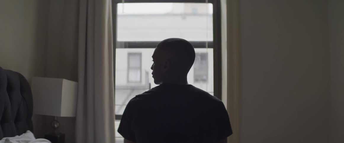 Sad looking male sitting on the side of the bed in front of a window staring at the headboard to the left of him