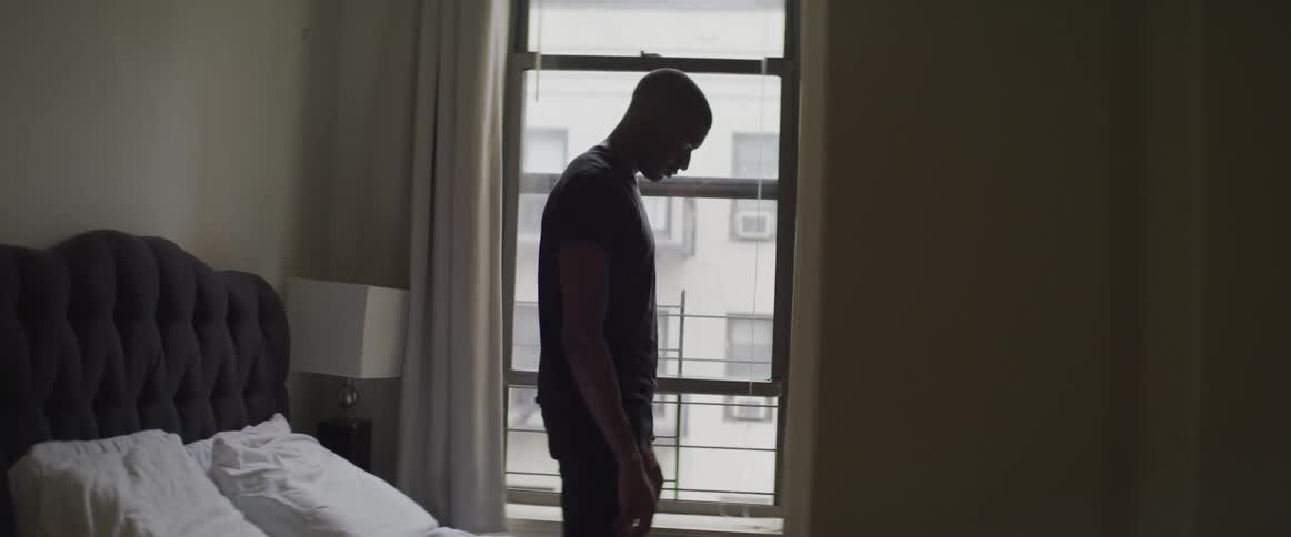 A man stands with his head down in his bedroom in front of a window