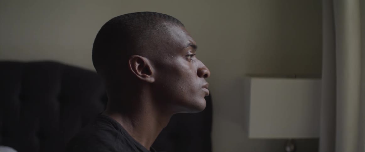A black man sits on a bed and raises his head and looks forwards