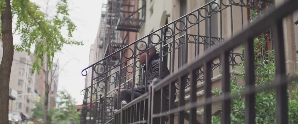 Male sitting on stairs outside of neighborhood building with his hands folded on his lap as he looks around