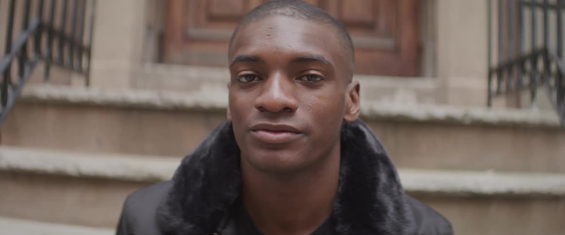A young black man sits on steps outside and turns his head and looks forwards