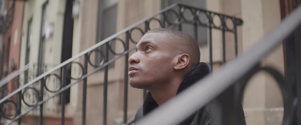 A black man in a coat sits on a neighborhood stoop
