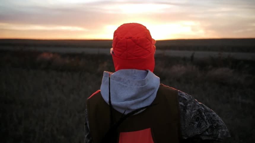 A man wearing an orange hat is walking on a field at sunset