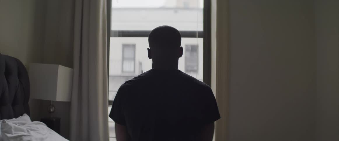 Male sits completely still on the side of the bed in front of the window in a bedroom