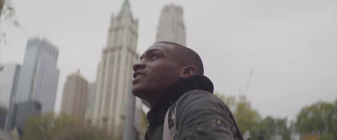A black man sits in a park in a city and looks into the distance