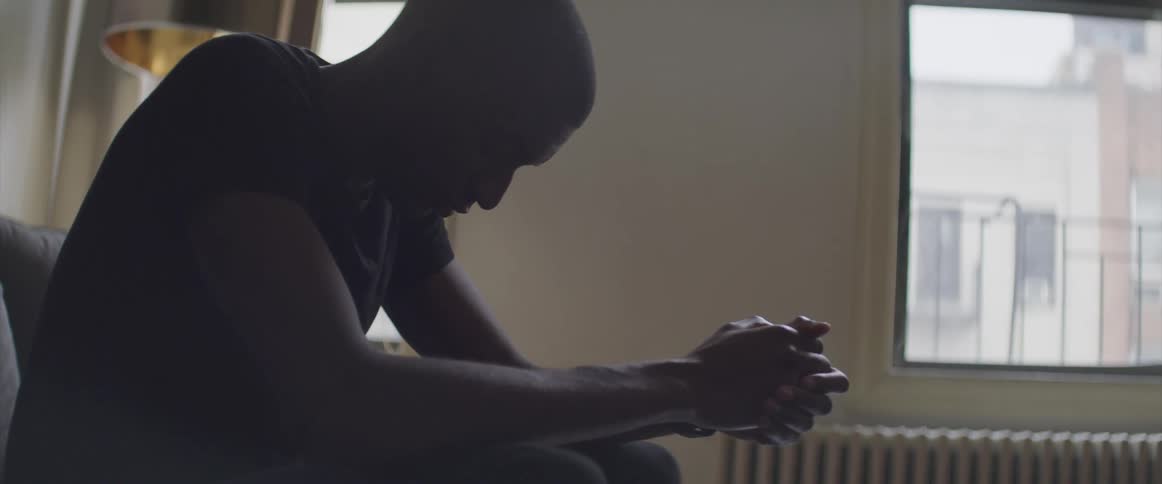 A man sits on the sofa in his living room with hands clasped in front of him and lowers his head