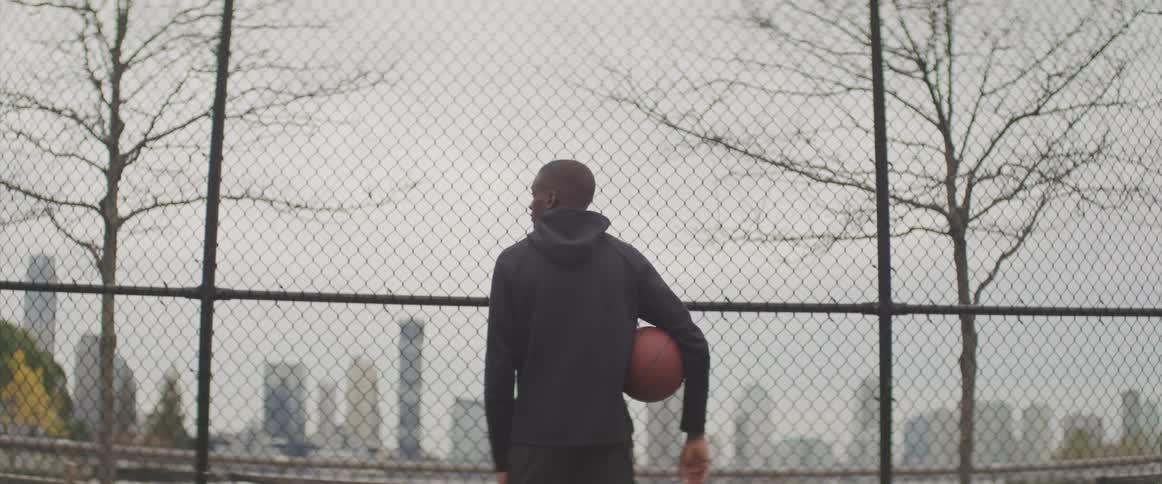 A man stands with a basketball under his arm in front of a chain link fence with views of a city skyline
