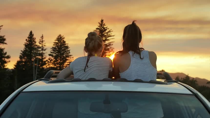 Two women watch a sunset standing in a car's sunroof opening