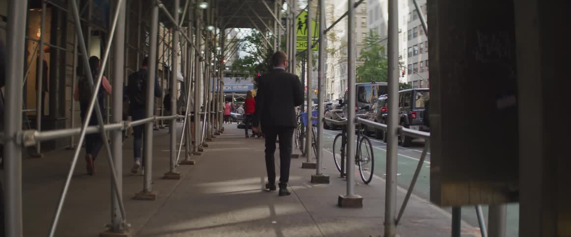A man walks a sidewalk under scaffolding in a city