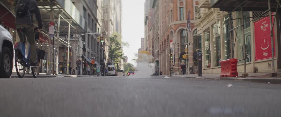 A man bikes along a city road as a few people walk the sidewalks