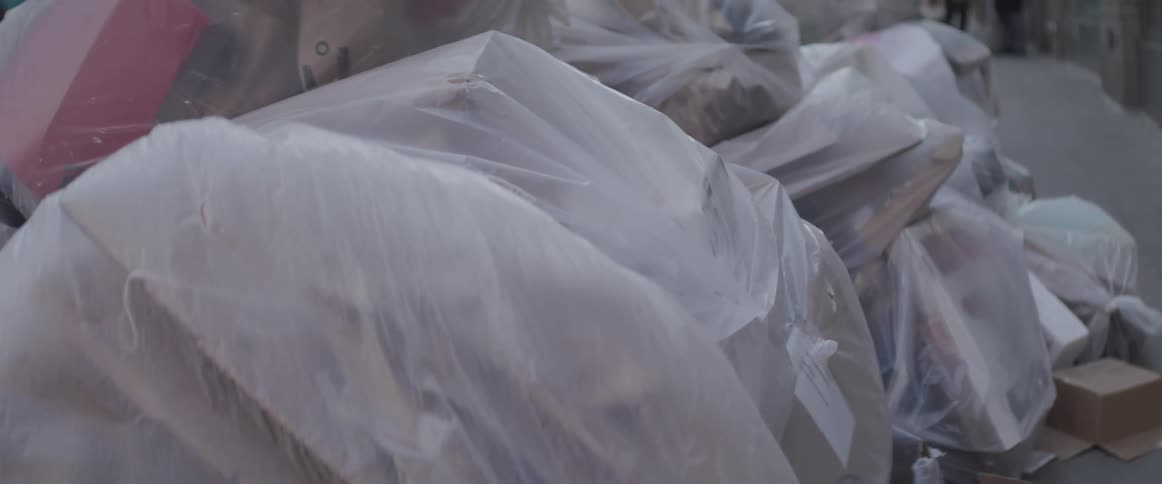 Bags of trash sit on a city sidewalk as pedestrians walk along