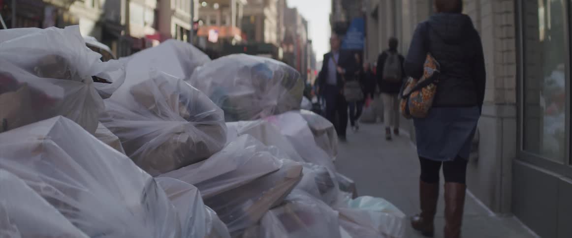 Pedestrians walking along a city sidewalk passing a big pile of trash bags thrown out on
